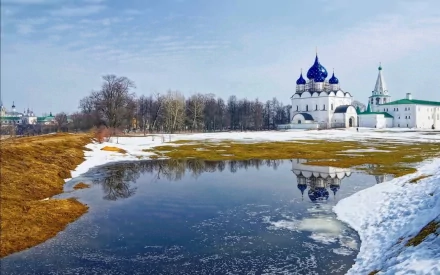 HD desktop wallpaper showcasing the Cathedral of the Nativity in Suzdal, reflected in a partially frozen river under a clear sky in early spring.