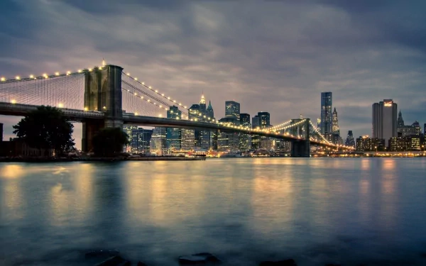 HD desktop wallpaper featuring the illuminated Brooklyn Bridge spanning over water with the Manhattan skyline in New York City under a moody evening sky.