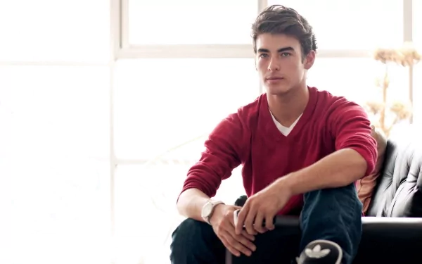 A man wearing a red shirt sits on a couch in a brightly lit room. The image is a beautiful HD desktop wallpaper and background.