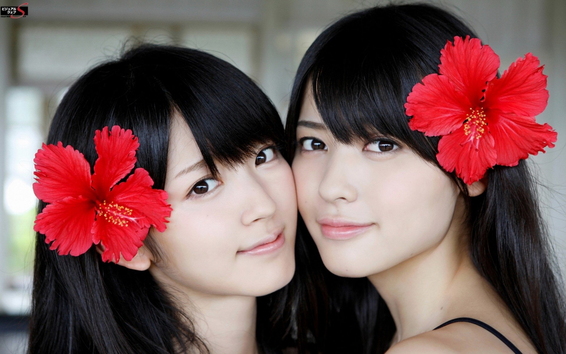 Two Asian women pose closely together, both wearing vibrant red hibiscus flowers in their hair, creating a lively and charming atmosphere in this HD desktop wallpaper.