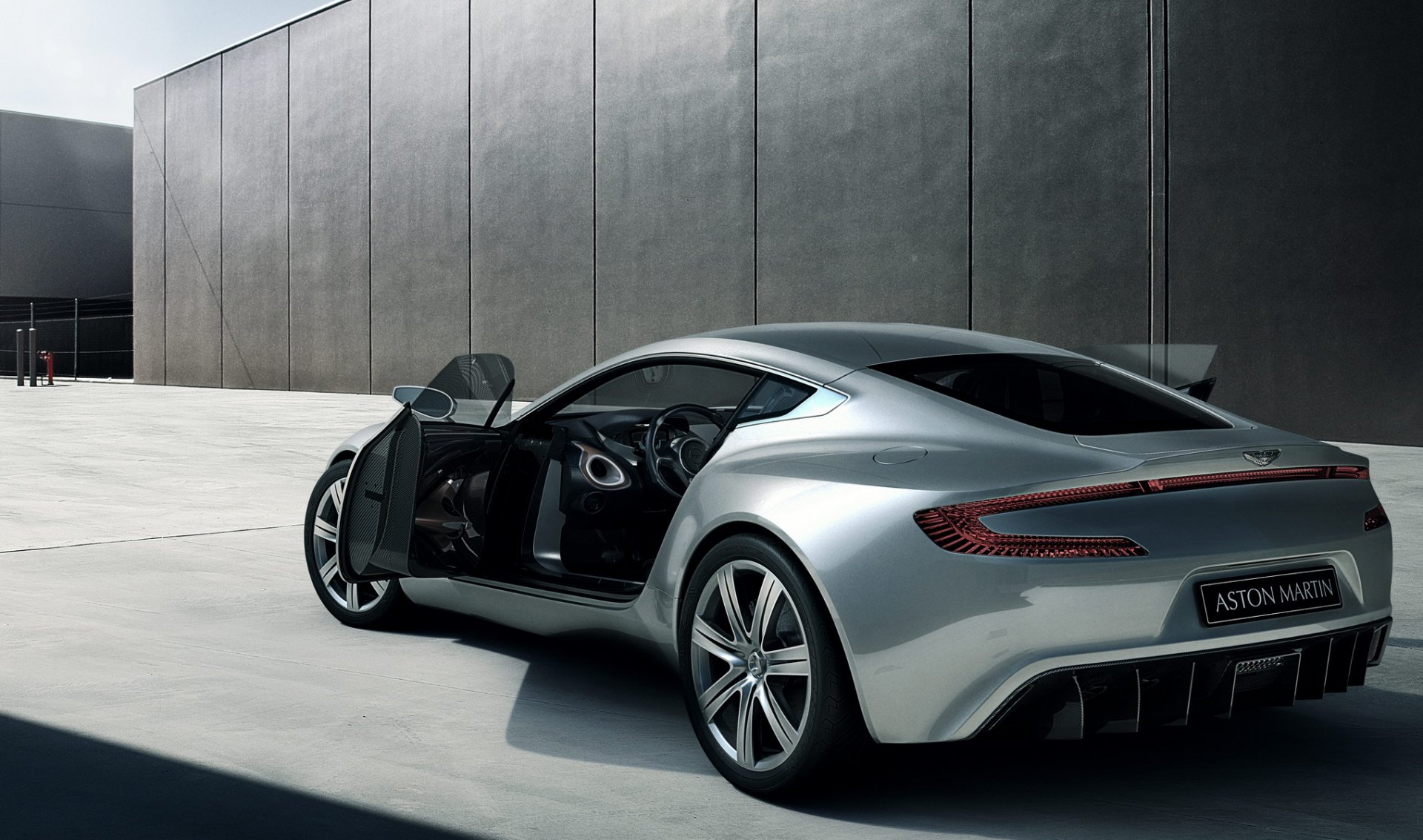 A sleek silver Aston Martin One-77 with its driver-side door open, set against a modern gray concrete wall, captured in an HD PC desktop wallpaper background.