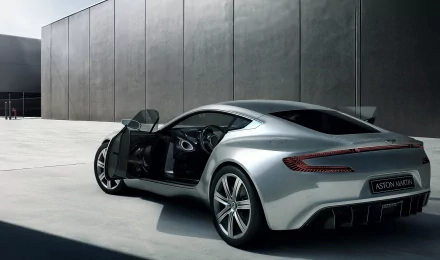 A sleek silver Aston Martin One-77 with its driver-side door open, set against a modern gray concrete wall, captured in an HD PC desktop wallpaper background.