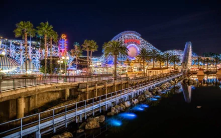 A vibrant amusement park at night, featuring colorful lights, palm trees, and a roller coaster reflected in calm waters, creating a captivating backdrop for any desktop wallpaper.