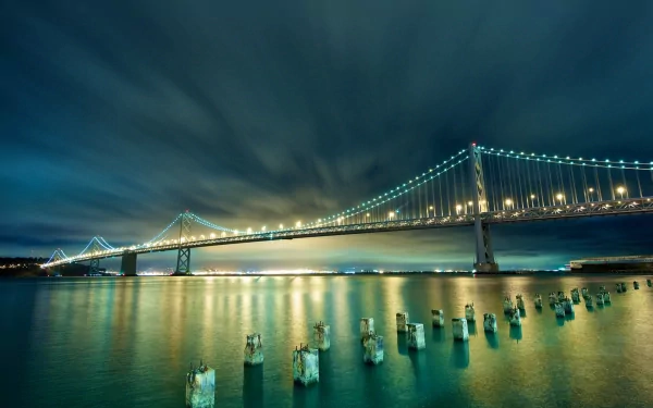 HD desktop wallpaper of San Francisco’s Bay Bridge illuminated at night, with calm waters reflecting the lights and old wooden pilings in the foreground.