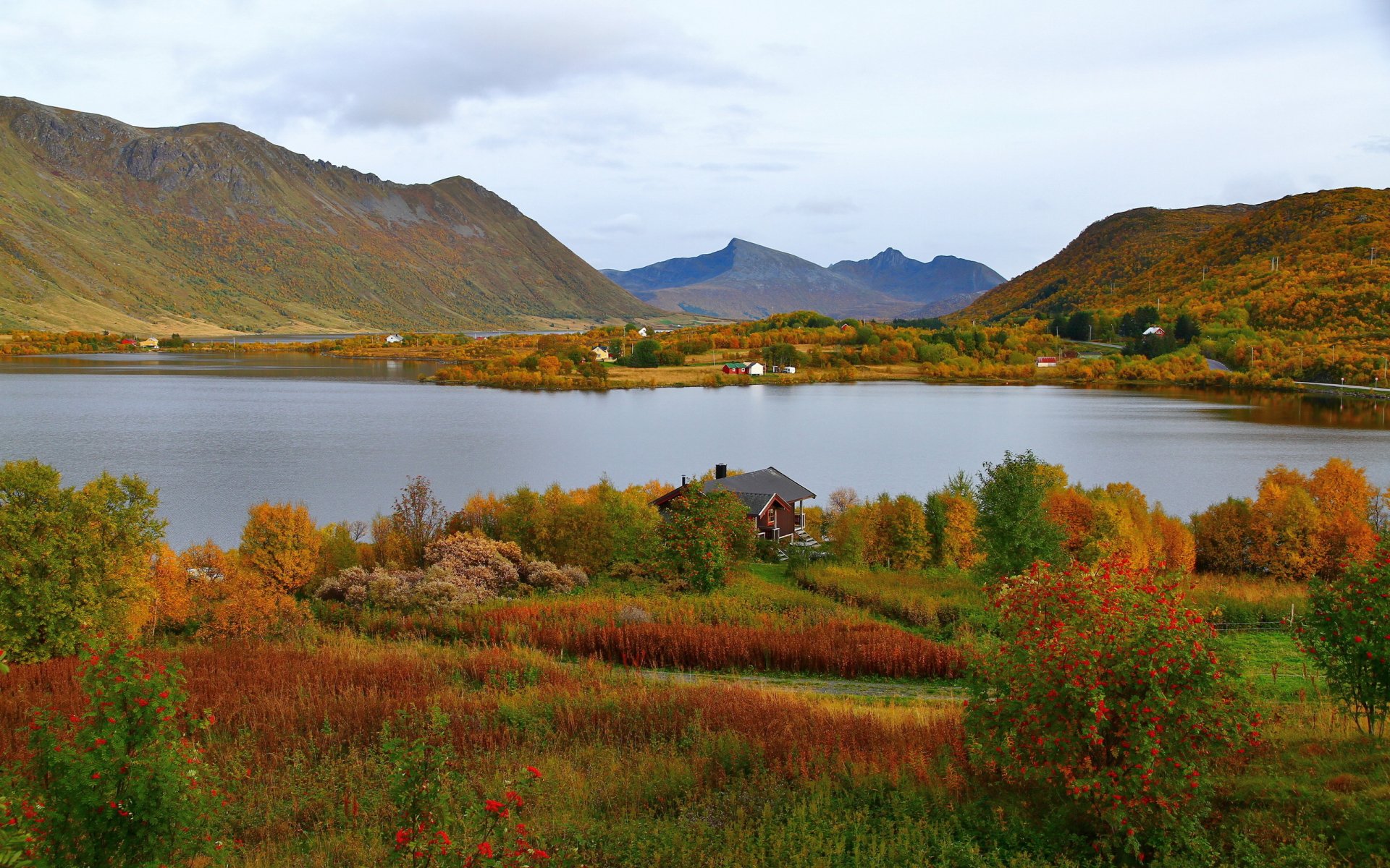 Photography, landscape — 2K Quad HD PC desktop wallpaper of an autumn lakeside valley with a small cabin, vivid fall foliage and distant blue-gray mountains beneath a soft, cloudy sky.
