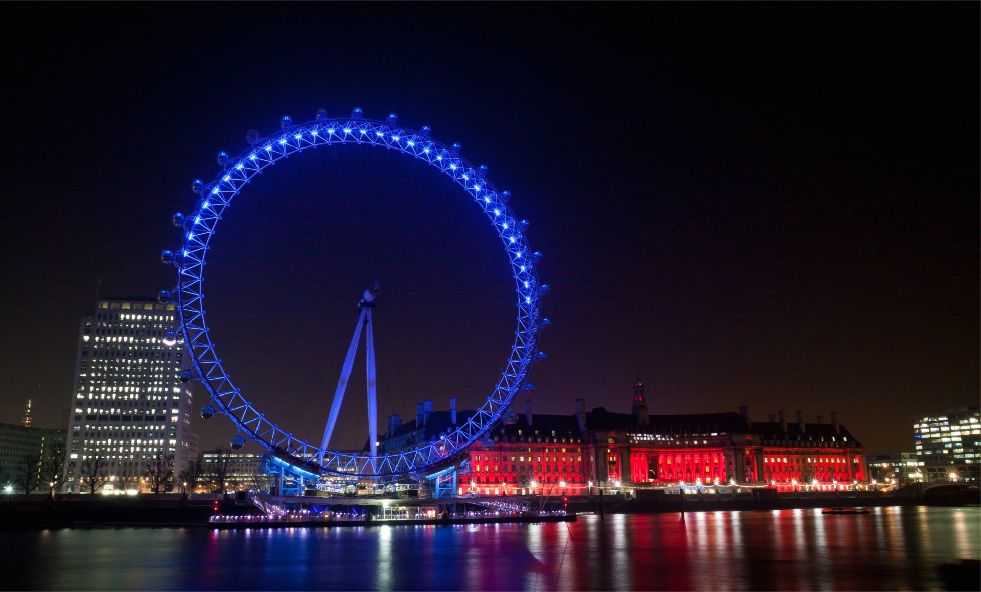 Night view of the London Eye ferris wheel in London, glowing blue above the Thames with illuminated riverside buildings; 4K Ultra HD desktop wallpaper of a man-made landmark.