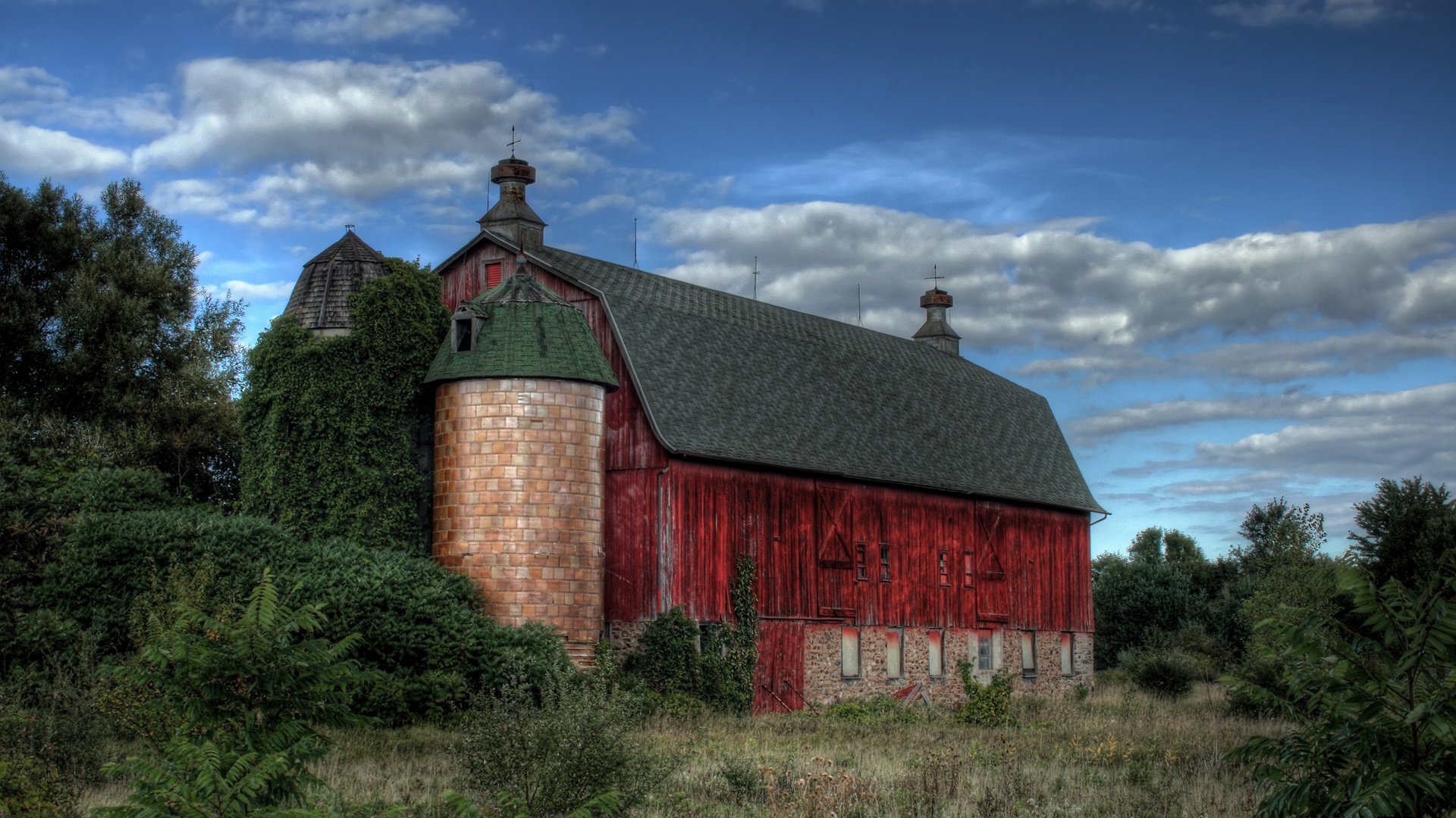 A vibrant red barn stands amidst lush greenery under a dramatic sky, showcasing a stunning man-made farm landscape. This HD image serves as an impressive desktop wallpaper.
