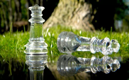 A close-up of two clear glass chess pieces set against a grassy background, reflecting in a surface, showcasing intricate details and a serene outdoor setting.