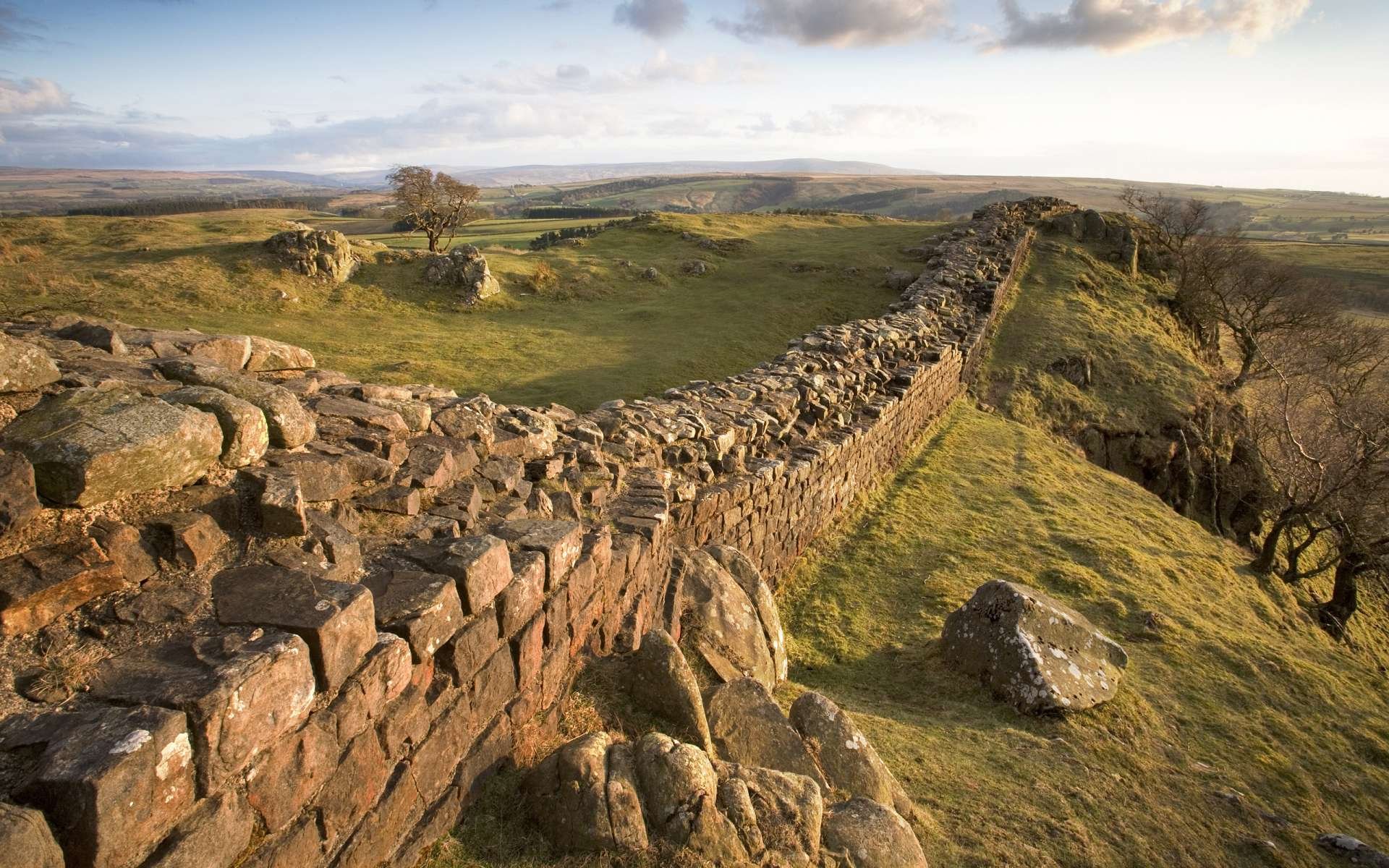 HD PC desktop wallpaper showcasing a scenic nature landscape with an ancient stone wall stretching across rolling green hills under a partly cloudy sky.