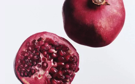 HD PC desktop wallpaper/background of a pomegranate food photo on white surface: one whole fruit and one halved, revealing glossy ruby seeds.
