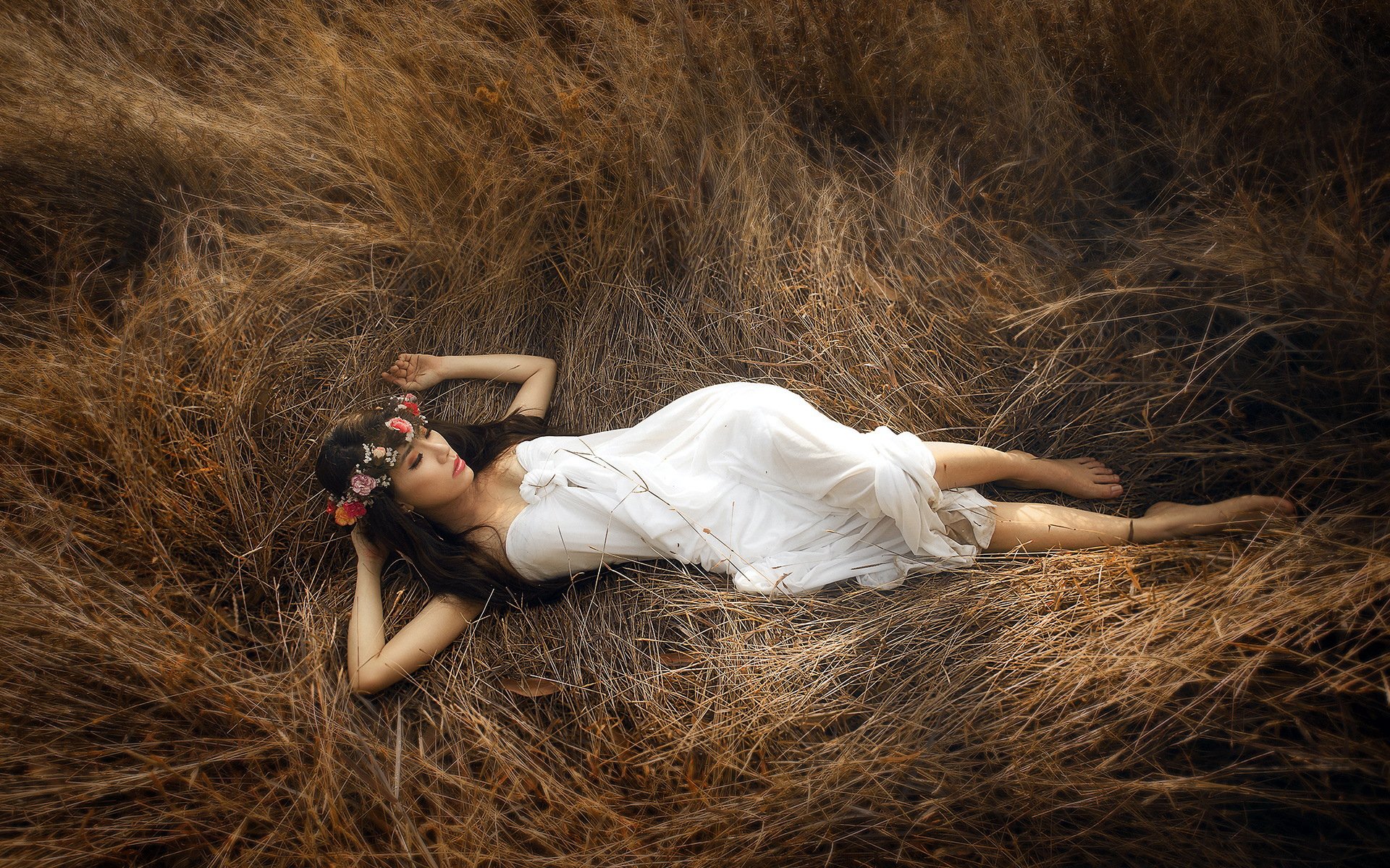A woman model in a white dress and flower crown lies in a field of dry grass, creating a serene and natural outdoor HD desktop wallpaper and background.