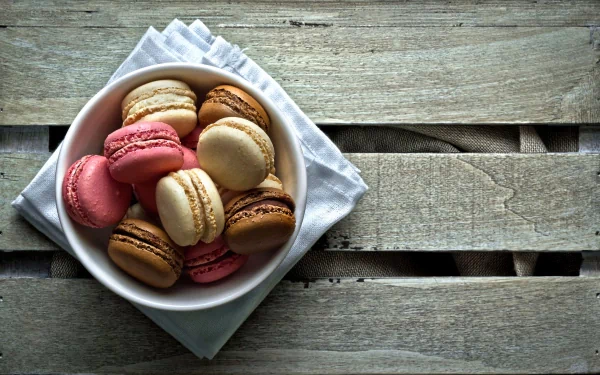 HD desktop wallpaper featuring a bowl of colorful macarons on a white napkin, set against a rustic wooden surface background.