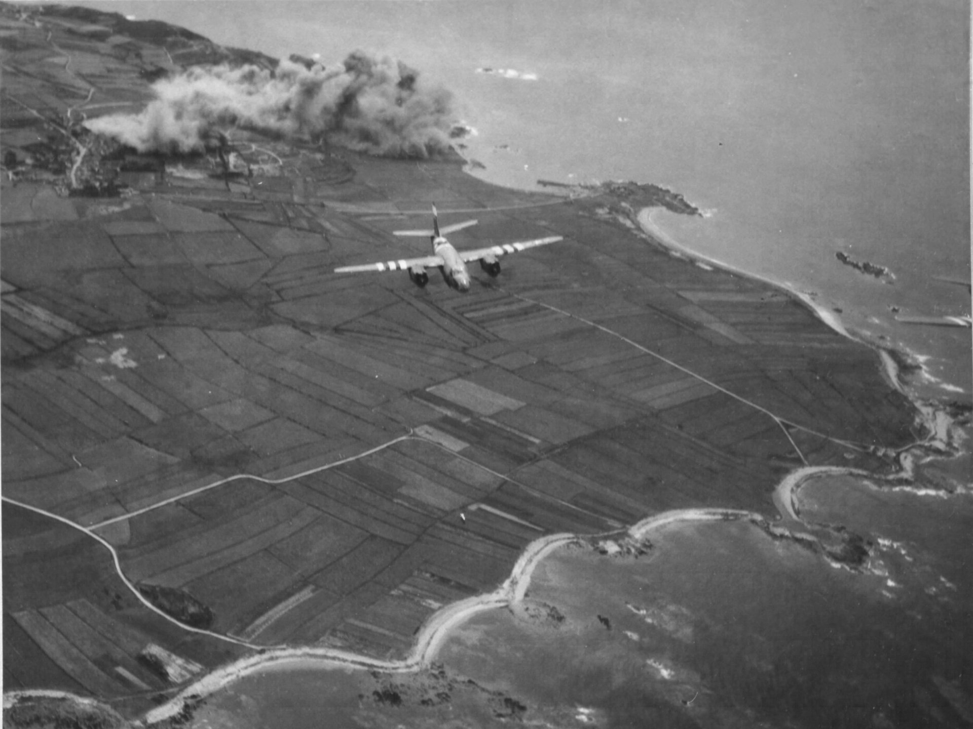 Grayscale 2K Quad HD PC desktop wallpaper showing a military transport aircraft flying over patchwork coastal fields and a winding shoreline.
