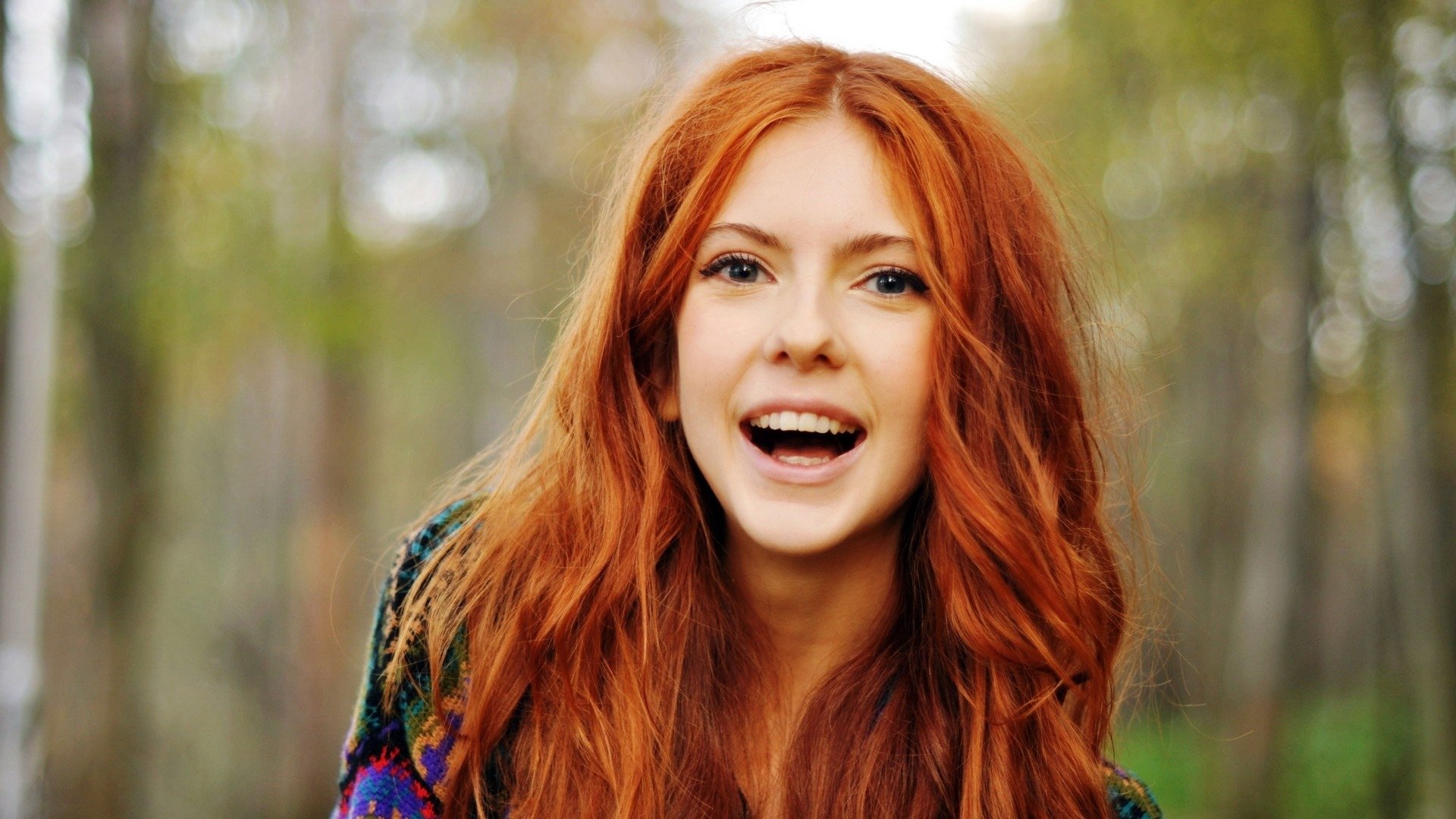 A vibrant HD desktop wallpaper featuring a smiling woman with long, wavy red hair, set against a natural background of soft greens and warm autumn colors.