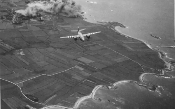 Grayscale 2K Quad HD PC desktop wallpaper showing a military transport aircraft flying over patchwork coastal fields and a winding shoreline.