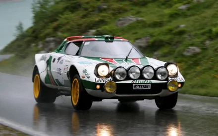 HD desktop wallpaper of a classic Lancia Stratos rally car speeding on a wet mountain road, showcasing its distinctive round headlights and vintage racing livery.