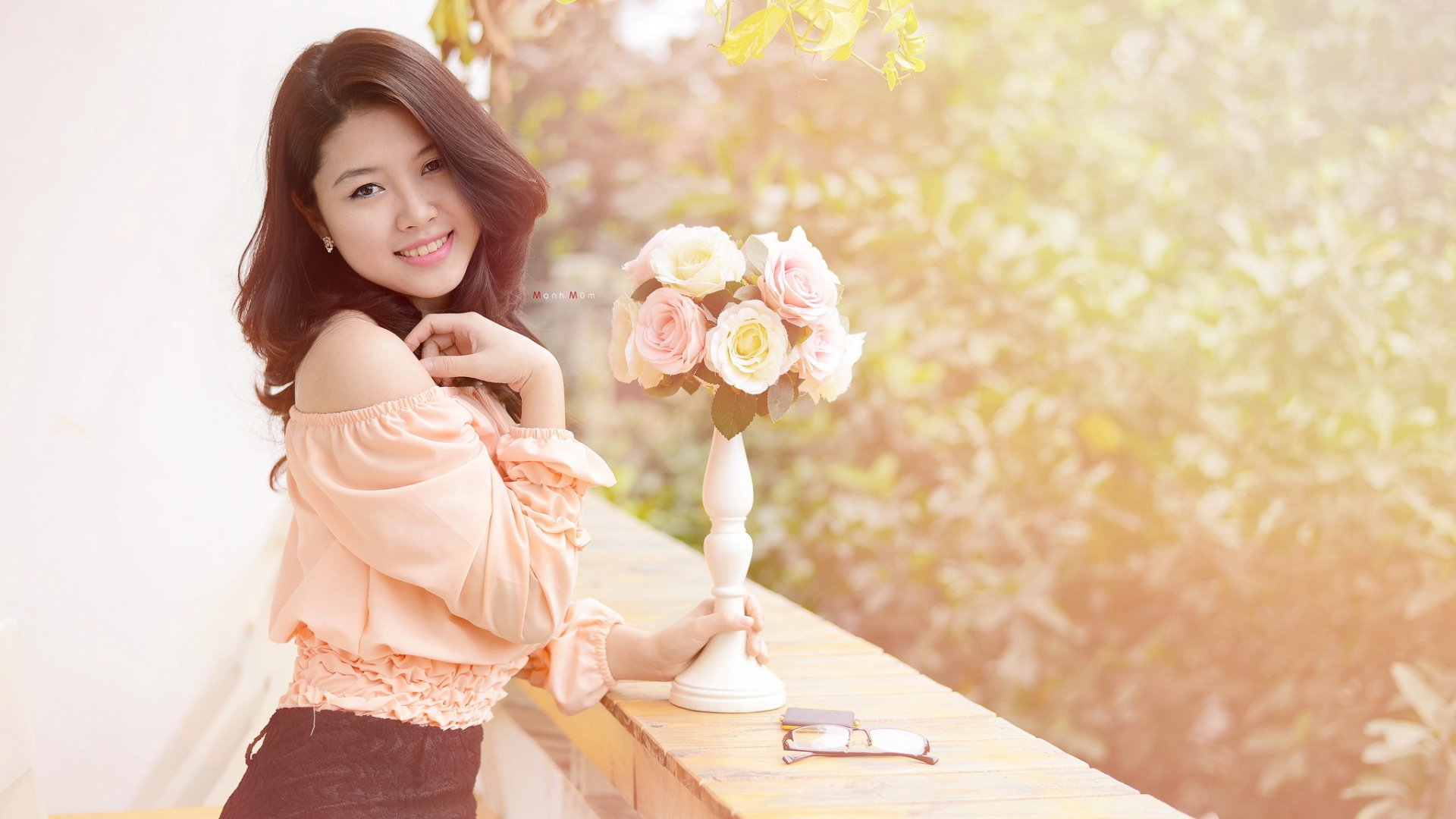 A smiling Asian woman poses outdoors beside a table with a vase of roses, set against a softly lit, lush green background. HD desktop wallpaper.