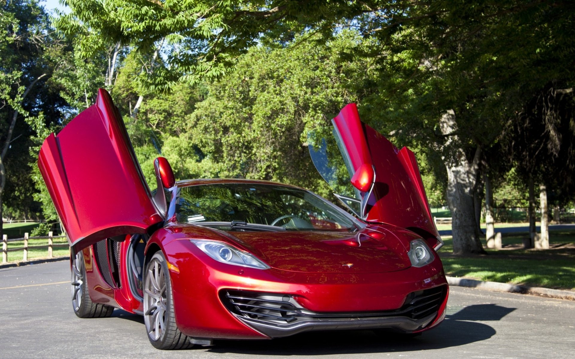 HD PC desktop wallpaper featuring a red McLaren sports car with its butterfly doors open, set against a leafy, sunlit backdrop.