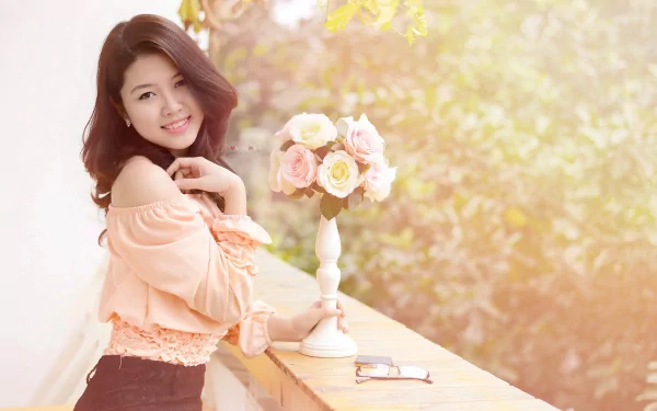 A smiling Asian woman poses outdoors beside a table with a vase of roses, set against a softly lit, lush green background. HD desktop wallpaper.