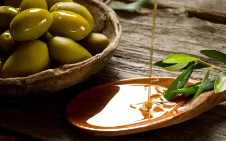 HD desktop wallpaper featuring green olives in a rustic bowl and golden olive oil pouring onto a wooden spoon, set on a textured wooden surface.
