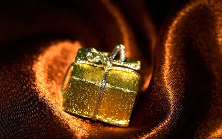 HD PC desktop wallpaper featuring a shimmering gold gift box on rich, textured brown fabric, capturing a warm and elegant festive atmosphere.