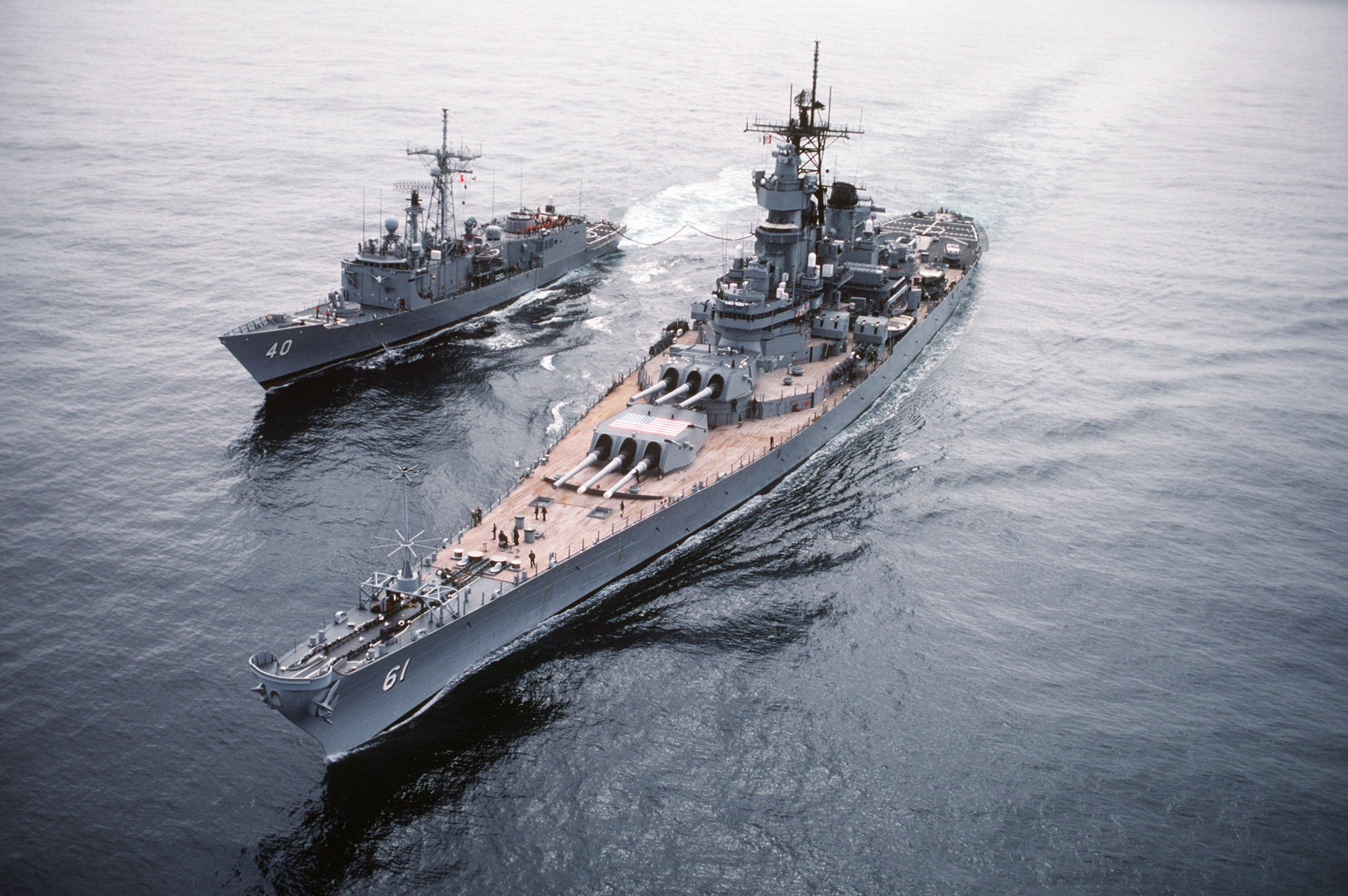 An aerial view of the USS Iowa (BB-61), a prominent battleship, accompanied by a smaller military vessel, showcasing naval power on the open sea as a striking HD desktop wallpaper.