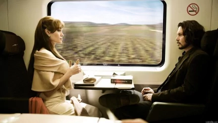 HD PC desktop wallpaper from the movie The Tourist: a woman in beige and a man in a dark coat sit facing each other in a train compartment, blurred fields passing by outside the window.