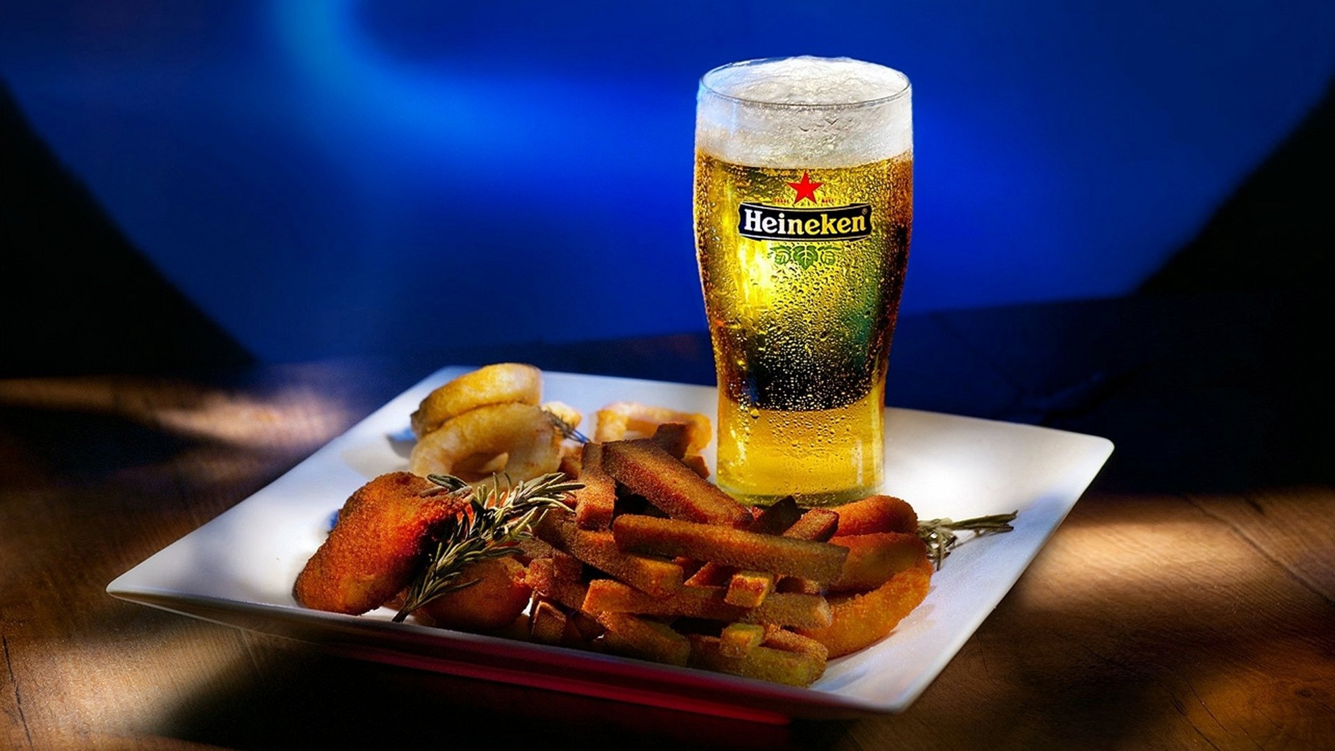 A vibrant HD desktop wallpaper featuring a pint of Heineken beer next to a plate of deliciously arranged food on a dark, blurred background.