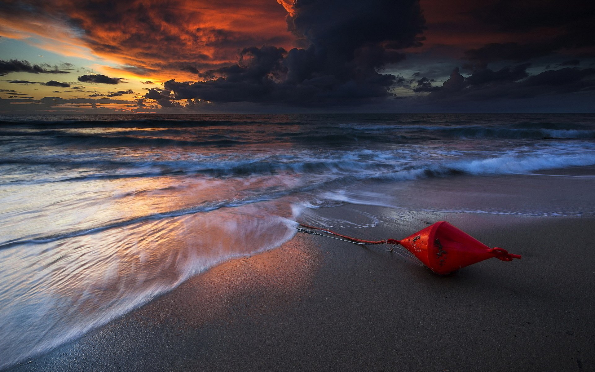 HD PC desktop wallpaper showcasing a scenic nature view of a dramatic sunset over ocean waves washing onto a sandy beach with a red buoy resting on the shore.