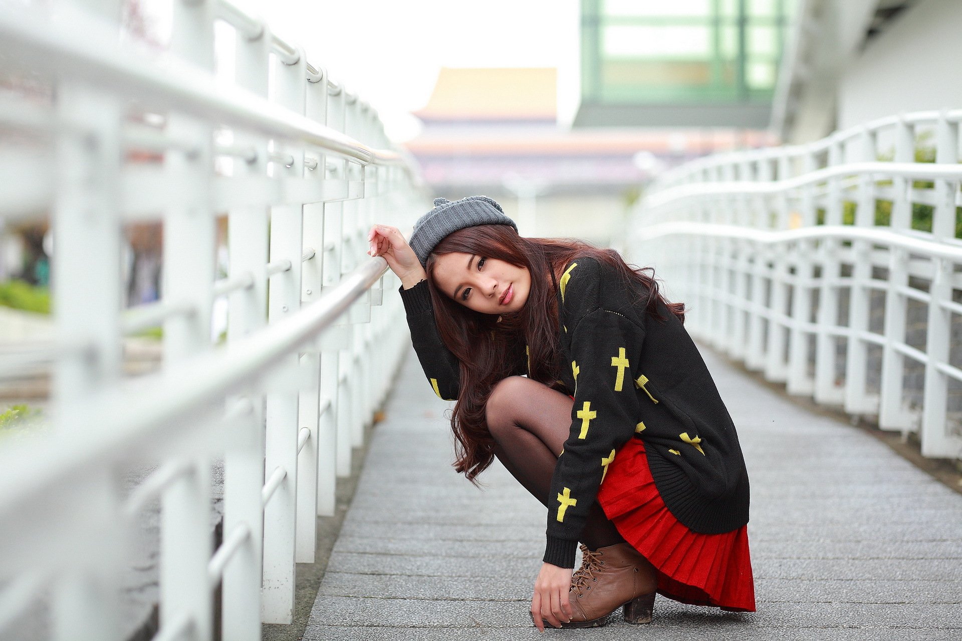 A woman in a stylish outfit with a beanie poses playfully on a walkway, surrounded by a modern architectural background. This HD image serves as an engaging desktop wallpaper.