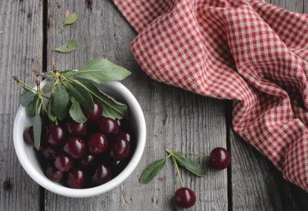 HD desktop wallpaper featuring fresh cherries with green leaves in a white bowl on rustic wooden surface next to a red checkered cloth.