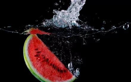 A vibrant slice of watermelon splashes into water against a black background, creating a striking HD desktop wallpaper showcasing refreshing summer vibes.