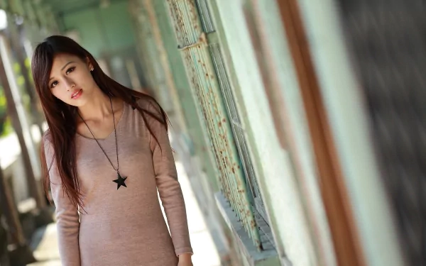 Asian woman with long hair stands outdoors near a green building, wearing a beige dress and a black star necklace. HD desktop wallpaper and background.