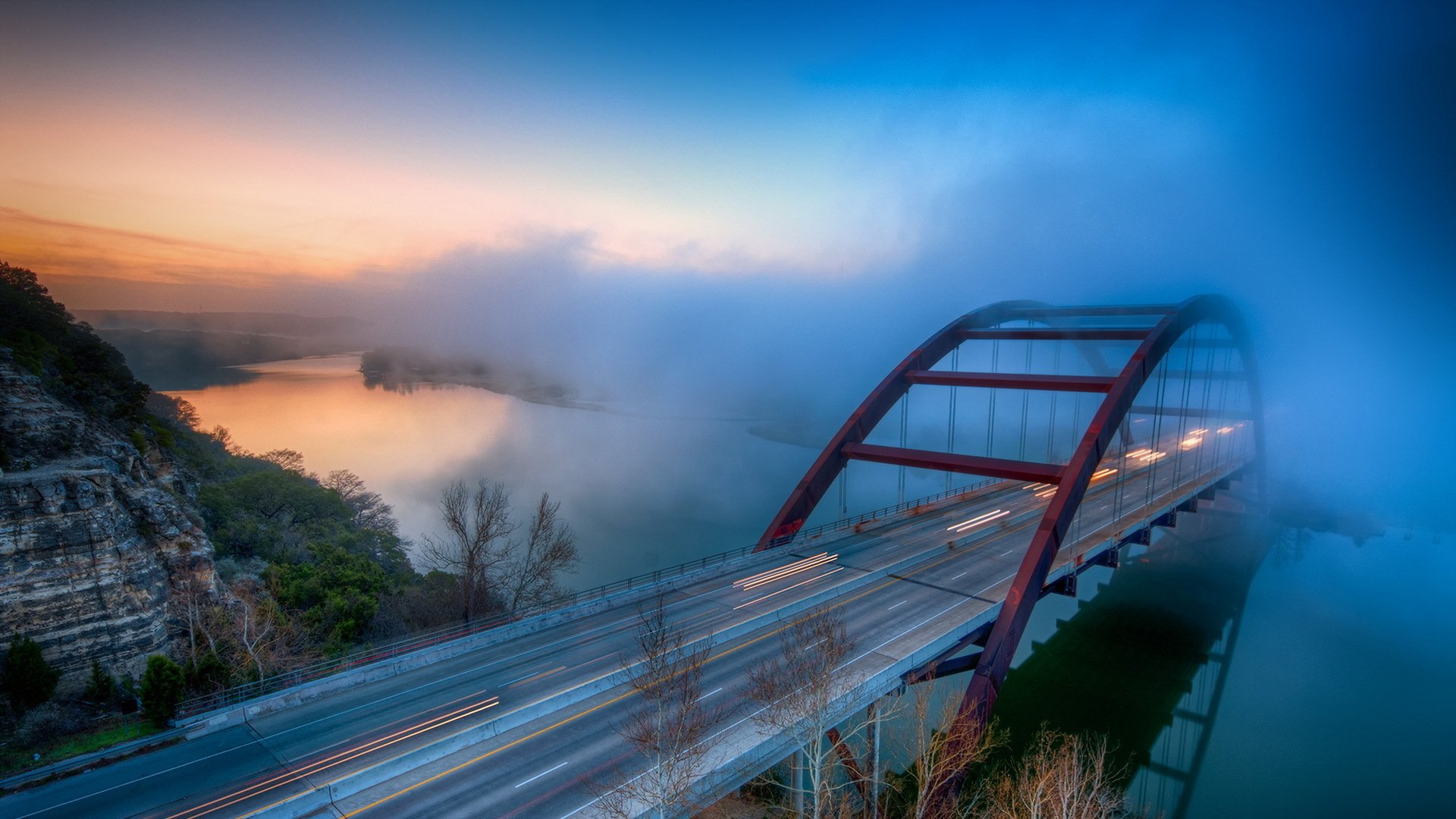 HD desktop wallpaper showing the Pennybacker Bridge in Austin, Texas, partially enveloped in mist during a vibrant sunrise or sunset.