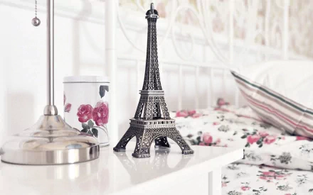 HD desktop wallpaper featuring a miniature Eiffel Tower on a bedside table, accompanied by a floral-patterned container and matching bedding, set in a cozy, softly lit bedroom scene.