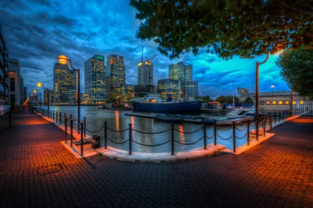 A stunning view of Canary Wharf in London, showcasing modern skyscrapers reflected in the water under a dramatic sky, with a charming waterfront promenade illuminated at dusk.