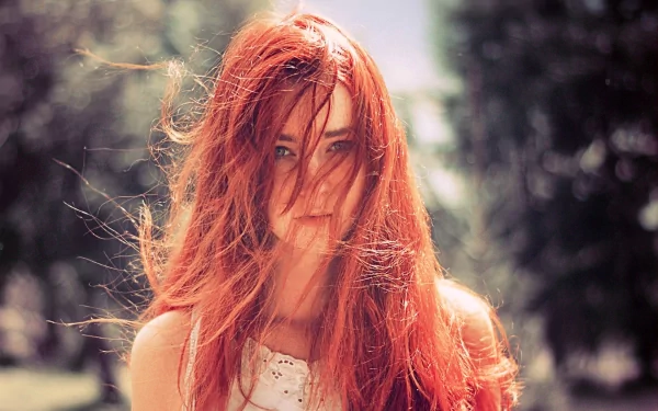 HD wallpaper of a woman with long, flowing red hair standing outdoors, her hair softly blowing in the wind. The background features blurred, lush green trees, creating a serene and natural ambiance.