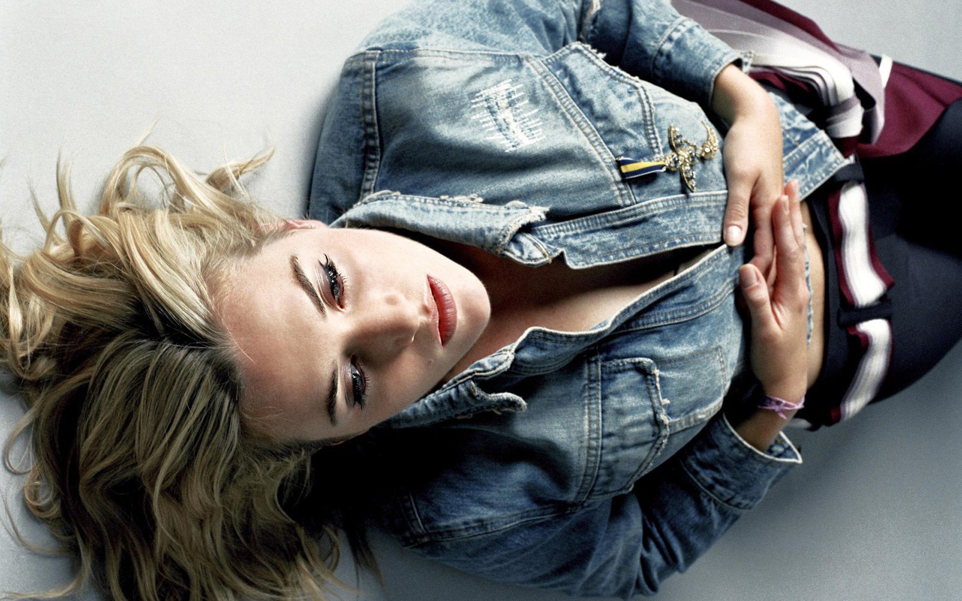 HD desktop wallpaper featuring celebrity Kate Winslet reclining with a thoughtful expression, wearing a denim jacket and dark pants against a neutral background.