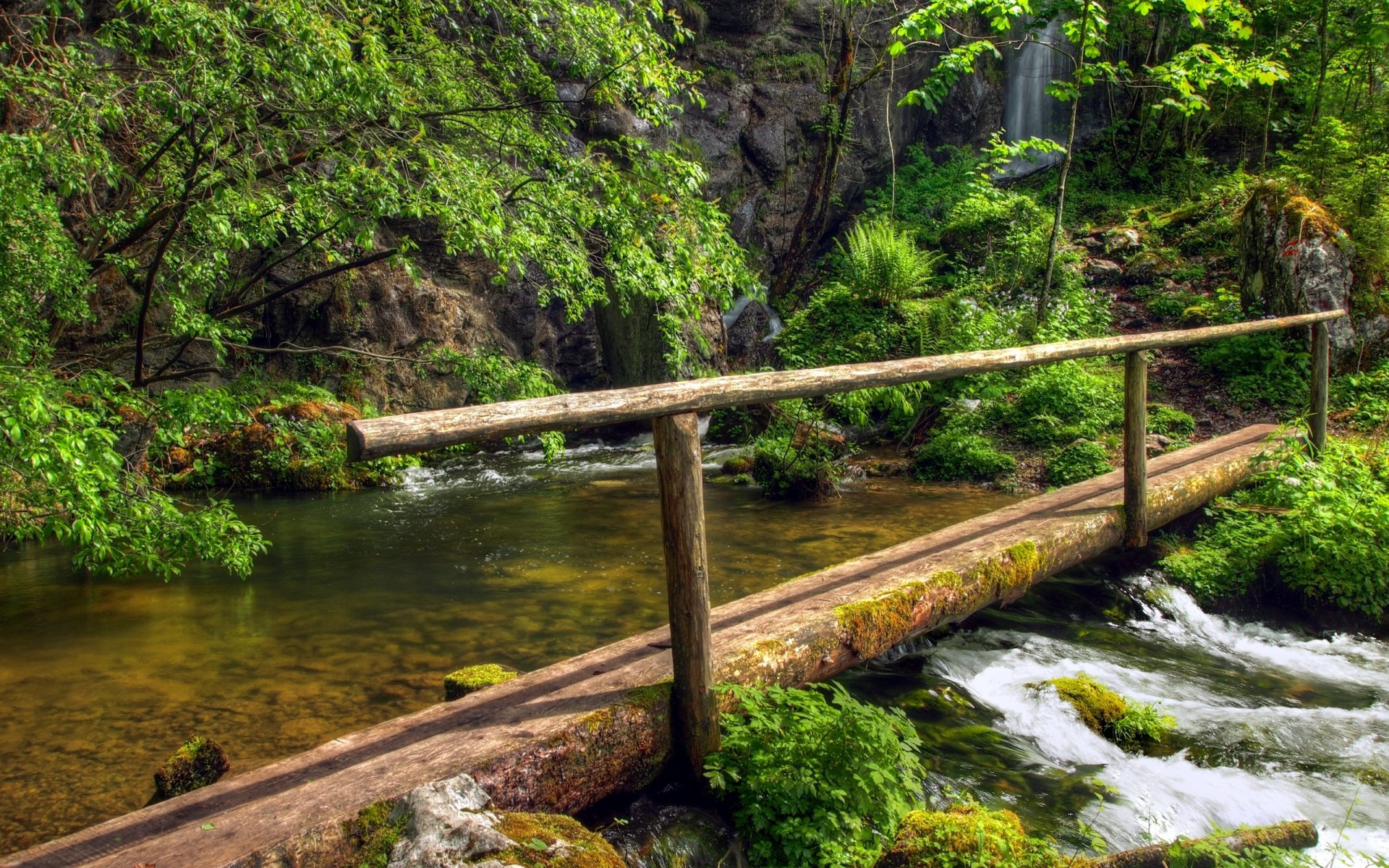 HD desktop wallpaper of a man-made wooden bridge crossing a clear stream surrounded by lush greenery and rocky terrain.