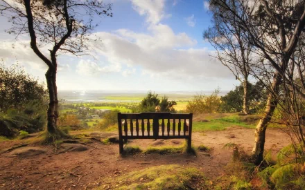 HD desktop wallpaper showing a man-made wooden bench overlooking a sunlit valley framed by trees under a partly cloudy sky.