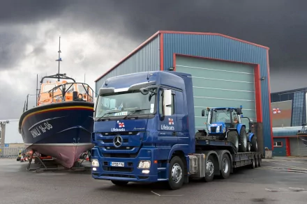 HD PC desktop wallpaper: blue Mercedes‑Benz Actros truck hauling equipment beside a lifeboat and marine shed under a dramatic, stormy sky.