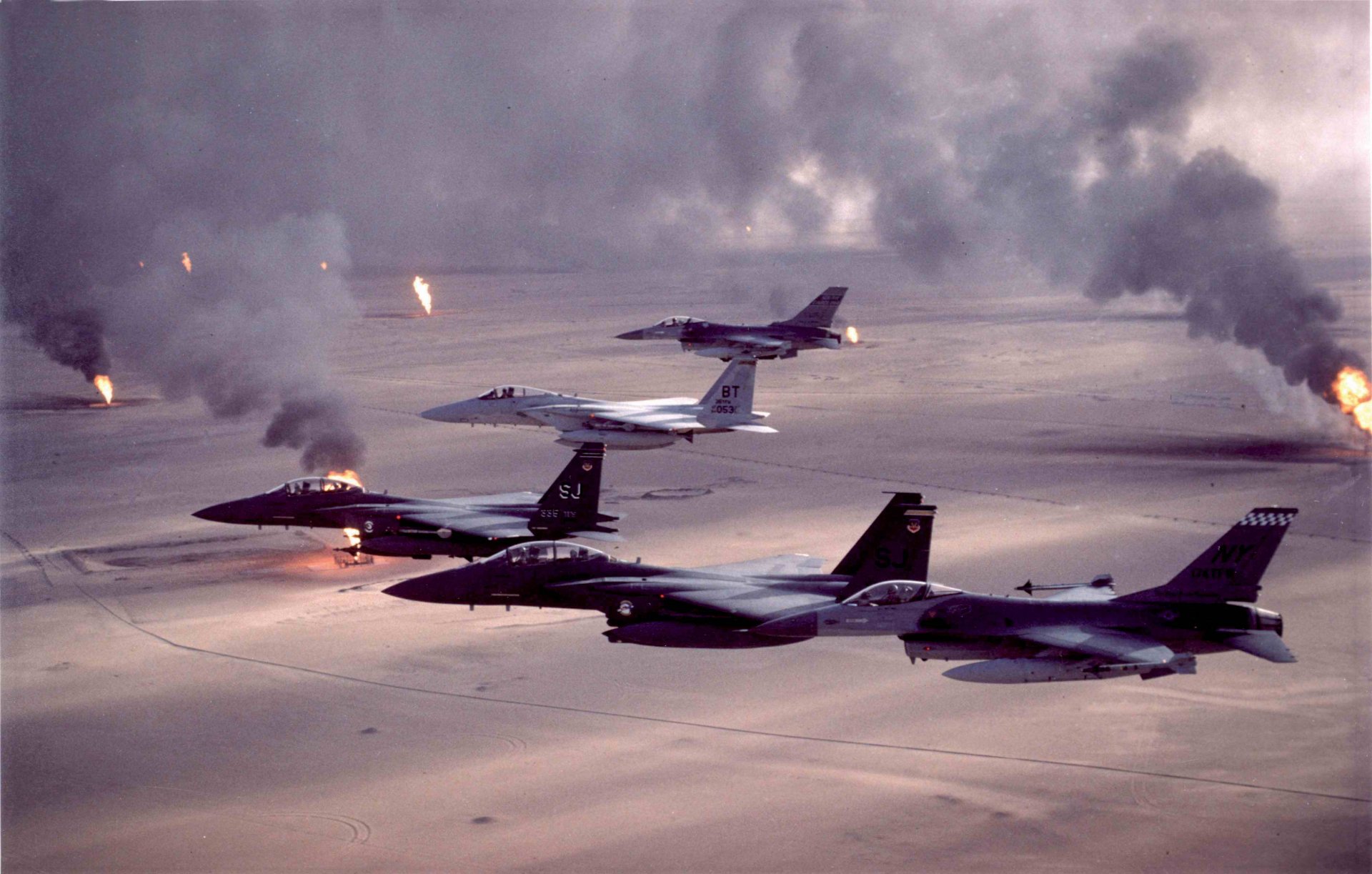 HD desktop wallpaper featuring military jet fighters in flight over a desert landscape, with smoke and fire in the background.