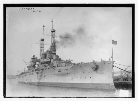 Black-and-white Ultra HD 5K PC desktop wallpaper of battleship USS Arkansas (BB-33) docked, smoke from funnels and US flag at stern — historic military warship profile.