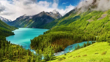 HD wallpaper featuring a stunning landscape with a turquoise lake, surrounded by lush green forests, majestic hills, and snow-capped mountains under a partly cloudy sky.