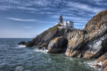 HD desktop wallpaper of a man-made lighthouse perched on rocky cliffs overlooking a calm sea under a partly cloudy sky.