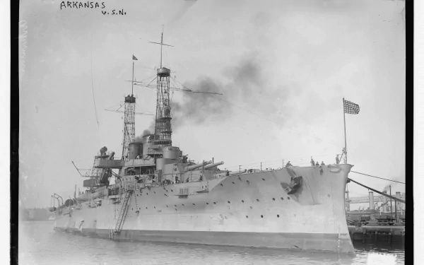 Black-and-white Ultra HD 5K PC desktop wallpaper of battleship USS Arkansas (BB-33) docked, smoke from funnels and US flag at stern — historic military warship profile.