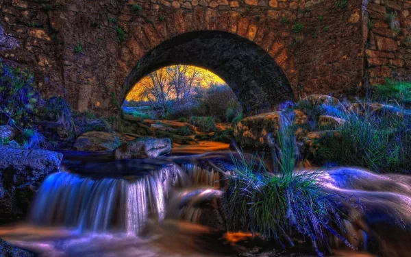 A serene HD desktop wallpaper featuring a stone bridge arching over a gently flowing stream, surrounded by lush greenery and illuminated by a colorful sunset.