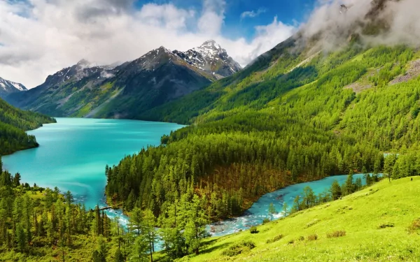 HD wallpaper featuring a stunning landscape with a turquoise lake, surrounded by lush green forests, majestic hills, and snow-capped mountains under a partly cloudy sky.