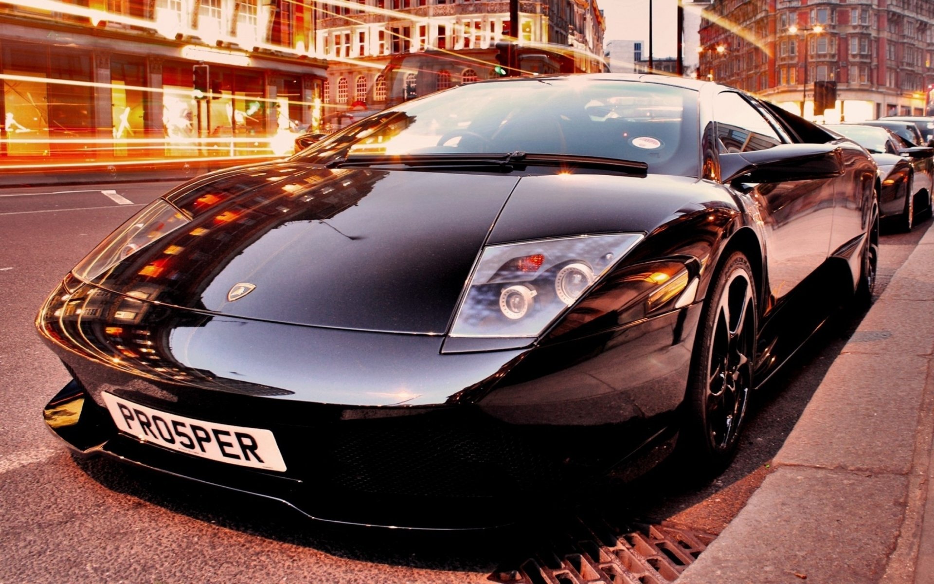 HD PC wallpaper featuring a sleek black Lamborghini Murcielago parked on a city street at dusk with motion blur from passing lights.