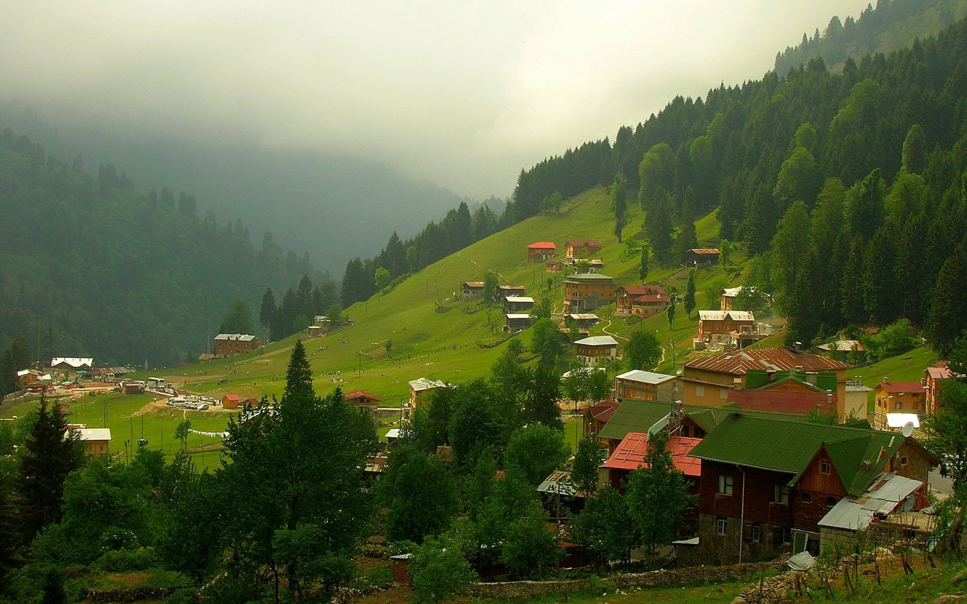 HD desktop wallpaper showing a serene man-made town nestled on green hills with surrounding forests and misty mountains in the background.